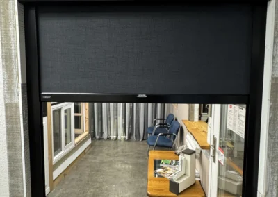 A black roller blind partially covers a window revealing a room with blue chairs, a wooden table, and a display case.