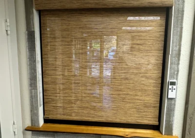 A window with a wooden ledge and a roller shade, framed in textured gray material, alongside a remote control.