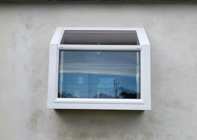 A modern white window with a slanted top, set in a textured beige wall, reflecting a cloudy sky and power lines.
